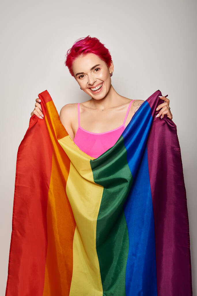 retrato de mulher feliz e jovem com cabelo rosa posando com bandeira do arco-íris lgbt em fundo cinza - Foto, Imagem