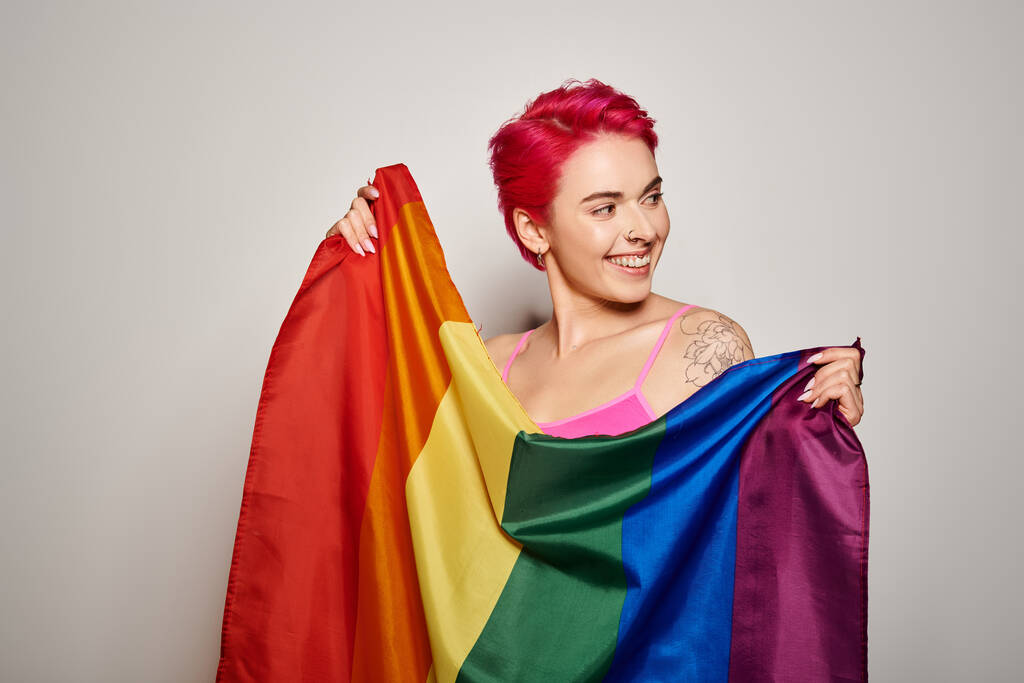 Porträt einer zufriedenen Aktivistin mit rosa Haaren, die mit lgbt-Regenbogenfahne vor grauem Hintergrund posiert - Foto, Bild
