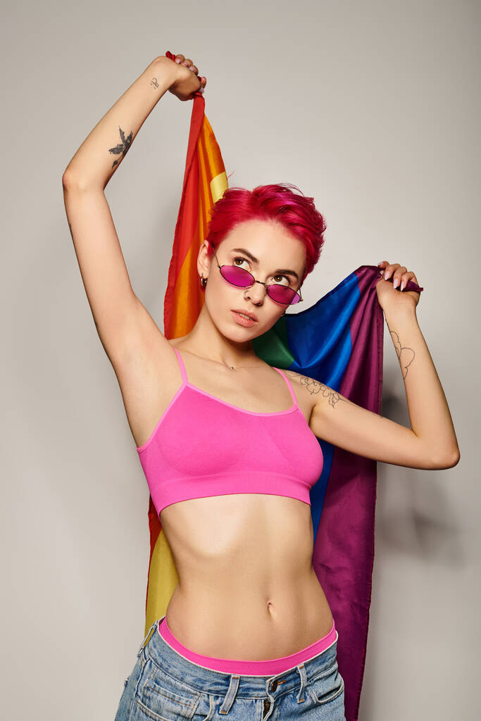 mujer joven tatuada con pelo rosa y gafas de sol posando con bandera de arco iris sobre fondo gris - Foto, Imagen