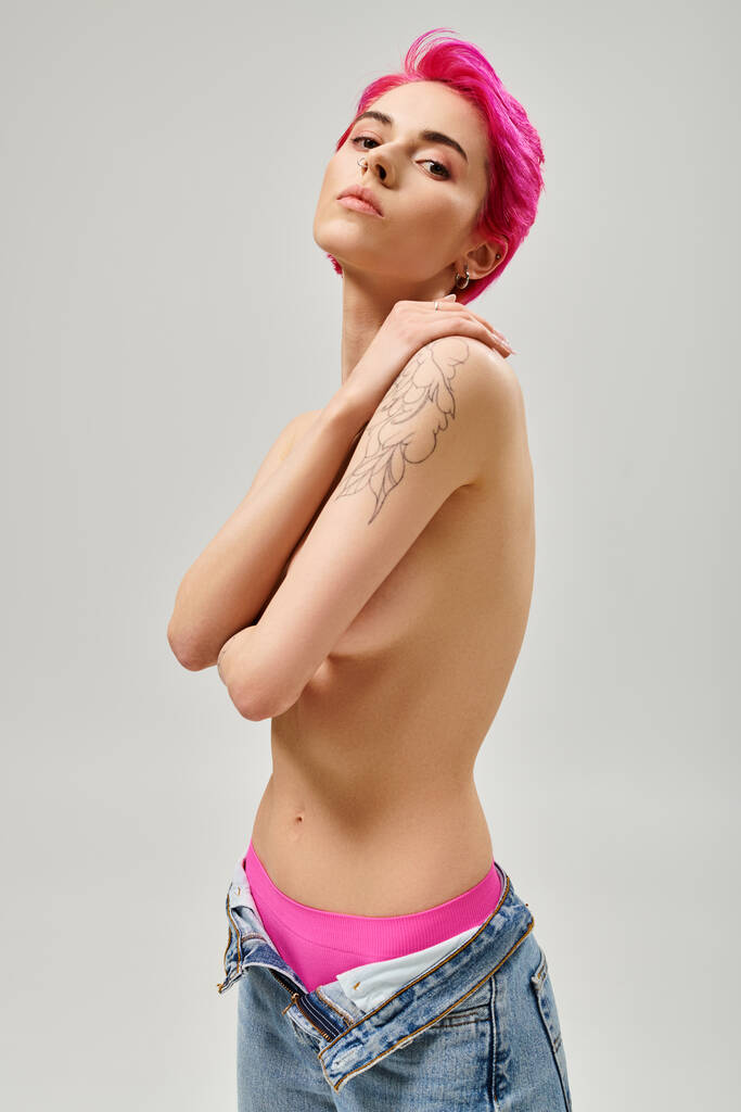 mujer joven tatuada y en topless con el pelo rosa cubriendo los pechos y posando sobre fondo gris - Foto, Imagen