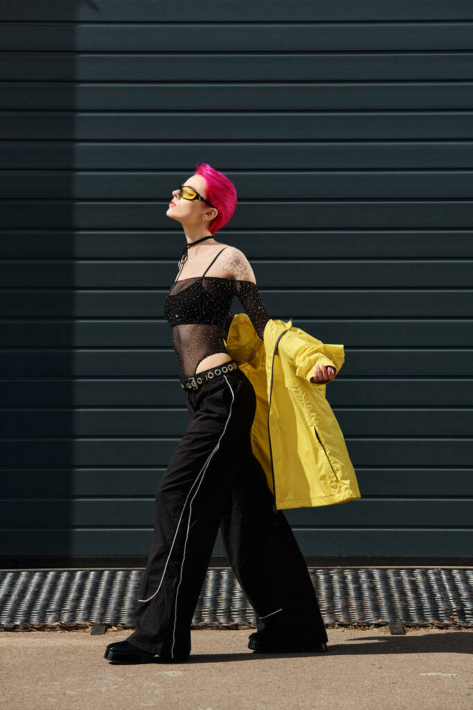 Pembe saçlı, sarı güneş gözlüklü ve modaya uygun giyinmiş genç bir kadın şehir caddesinde yürüyor. - Fotoğraf, Görsel