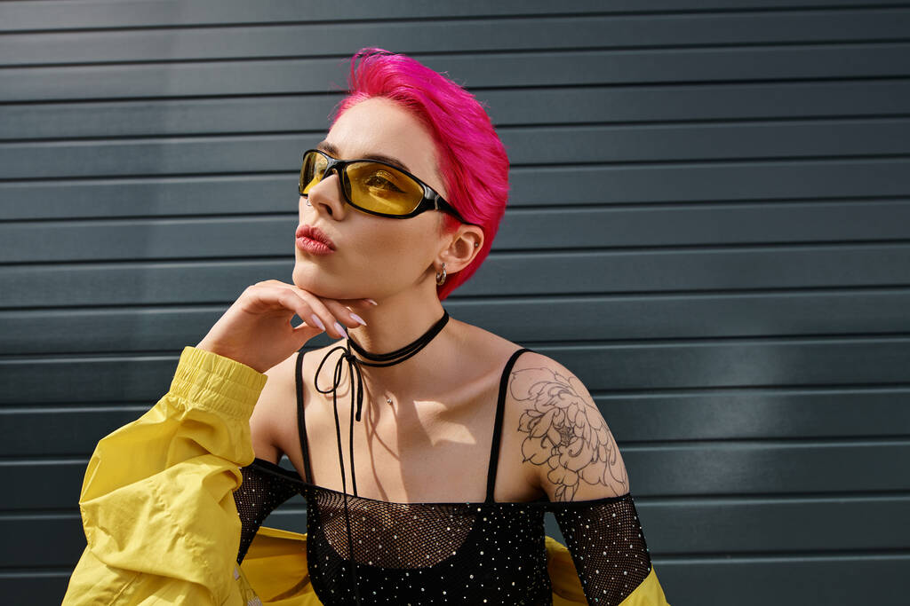 pensive young woman with pink hair and tattoo posing in sunglasses and trendy streetwear on street - Photo, Image