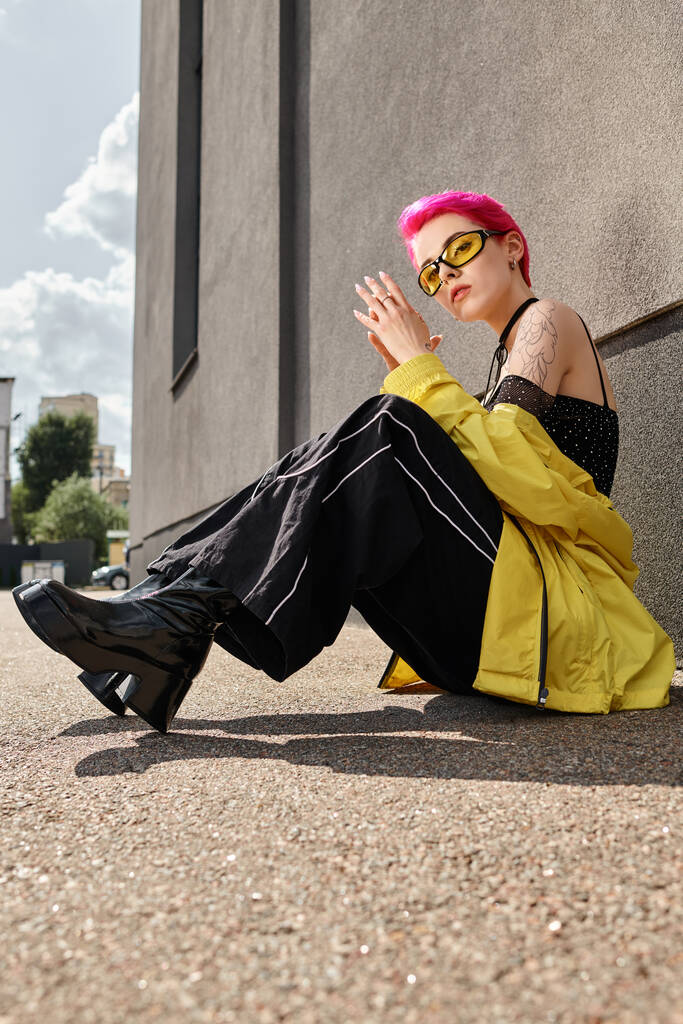 mulher de cabelos cor-de-rosa em óculos de sol amarelos e roupa da moda olhando para a câmera e posando na rua - Foto, Imagem