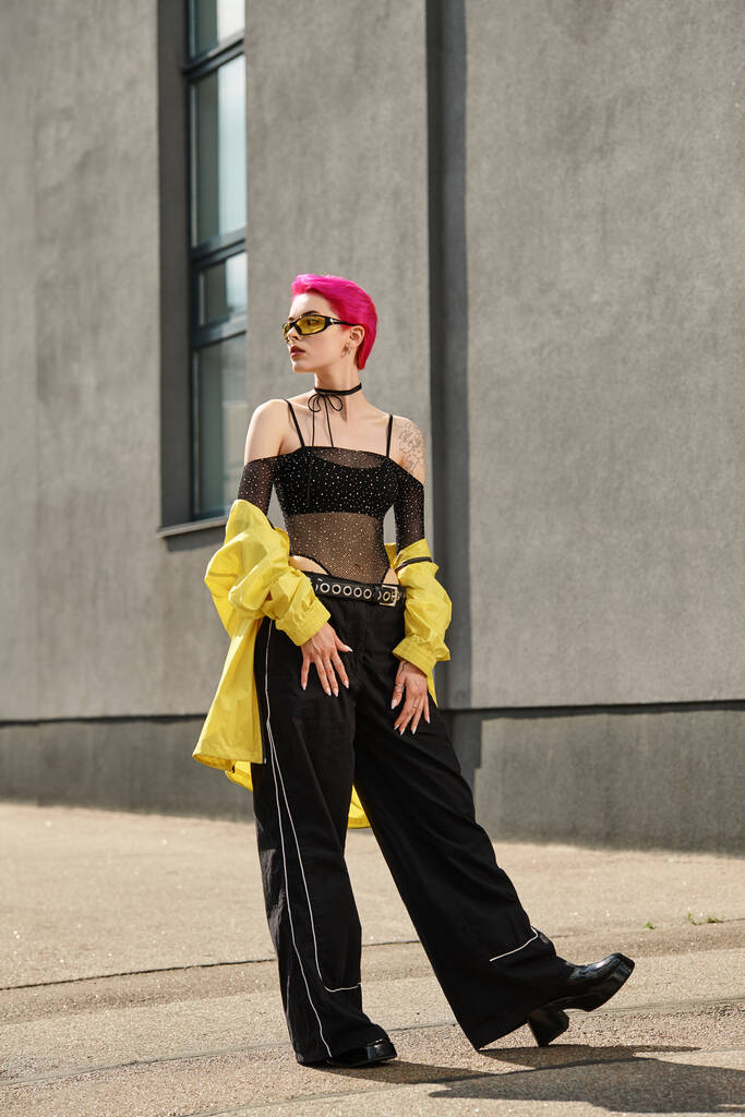 pink haired woman in yellow sunglasses and trendy outfit posing near building on urban street - Photo, Image