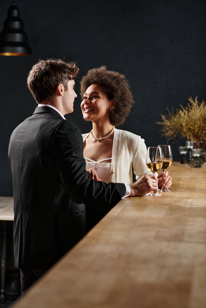 Χαρούμενη Αφροαμερικανή κυρία που κρατάει ποτήρι κρασιού και κοιτάζει τον άντρα κατά τη διάρκεια του ραντεβού την ημέρα του Αγίου Βαλεντίνου. - Φωτογραφία, εικόνα