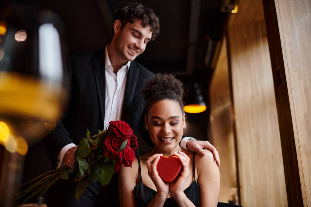 Mann hält am Valentinstag rote Rosen in der Nähe einer jungen afrikanisch-amerikanischen Frau mit herzförmiger Schachtel - Foto, Bild