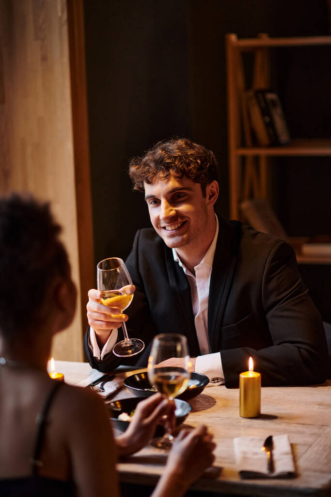 radostný muž v elegantním oblečení při pohledu na přítelkyni se sklenkou vína během rande v restauraci - Fotografie, Obrázek