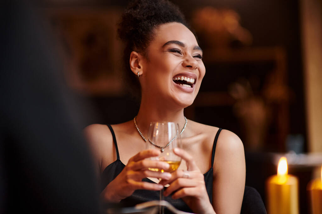 χαρούμενη Αφροαμερικανή γυναίκα που γελάει και κρατάει ένα ποτήρι κρασί κατά τη διάρκεια του ραντεβού την ημέρα του Αγίου Βαλεντίνου - Φωτογραφία, εικόνα