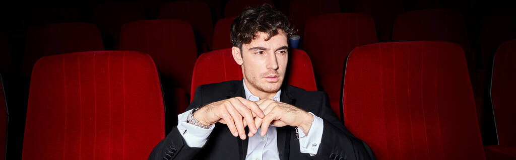 przystojny elegancki mężczyzna z eleganckim stylu siedzi na czerwonym kinie krzesła i patrząc daleko, sztandar - Zdjęcie, obraz