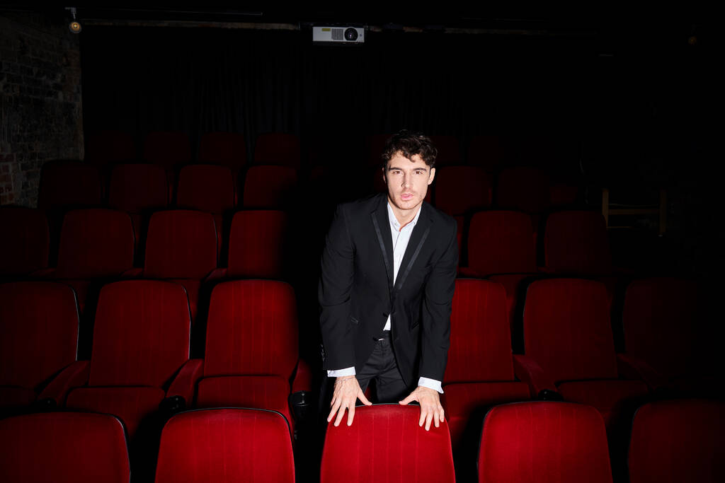 красивый молодой человек в черном элегантном костюме позирует между красными стульями кинотеатра и смотрит в камеру - Фото, изображение