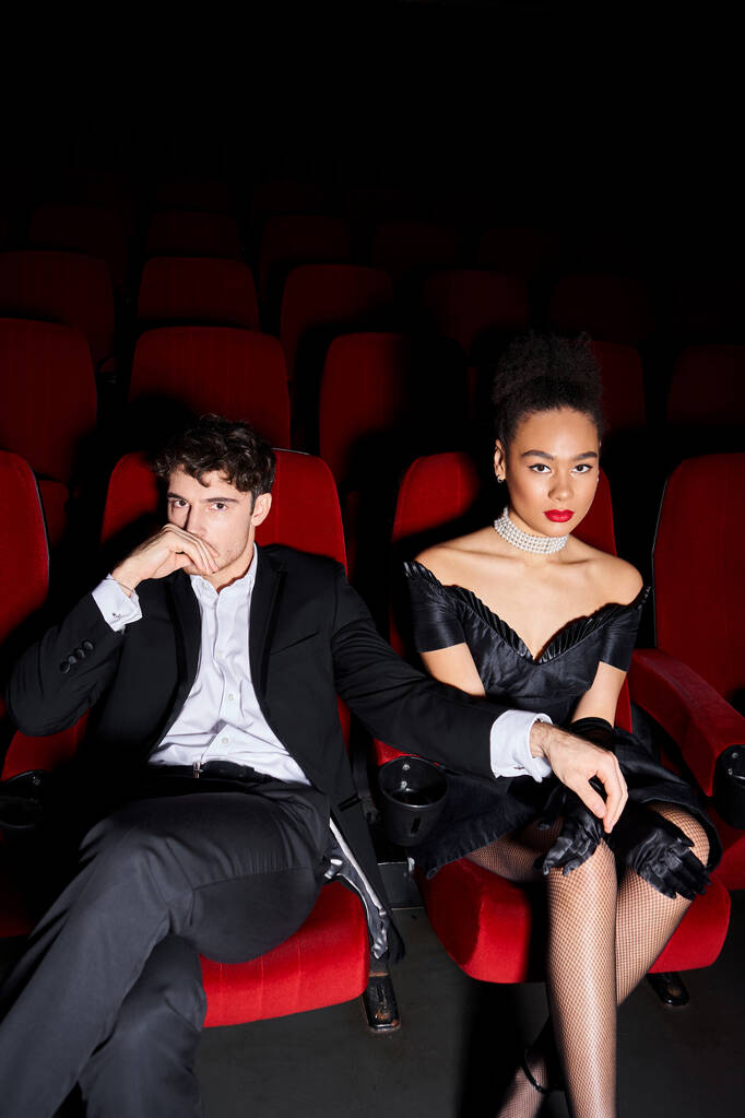 attraente coppia multiculturale in posa insieme su sedie cinema rosso e guardando la fotocamera in data - Foto, immagini