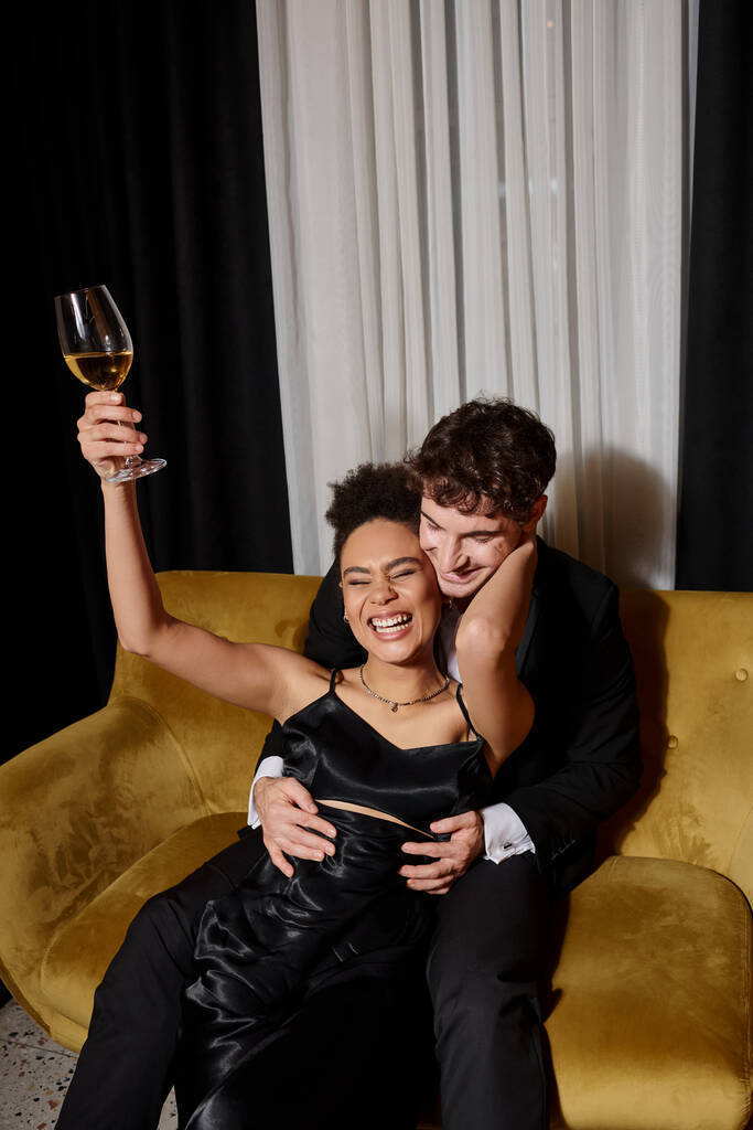 happy african american woman holding glass of wine and laughing near man in suit on velvet sofa - Photo, Image