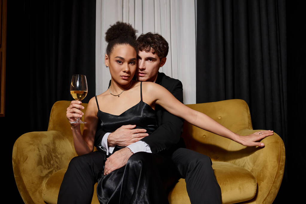 uomo abbracciando elegante donna afro-americana in possesso di un bicchiere di vino e seduto su un divano di velluto - Foto, immagini
