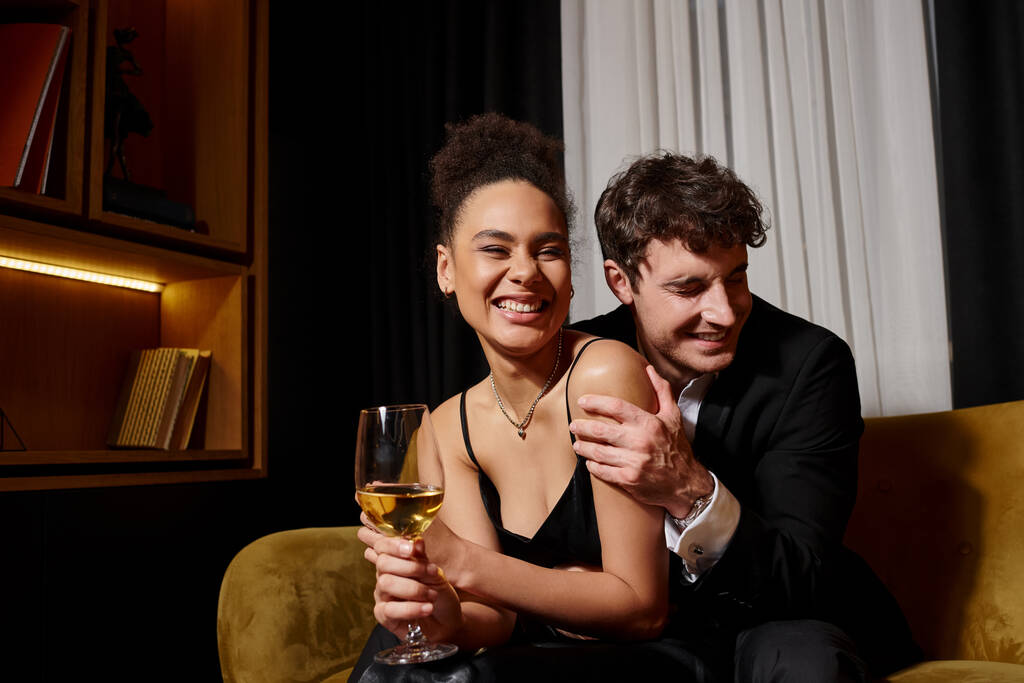 man hugging excited african american woman holding glass of wine and sitting on velvet sofa - Photo, Image