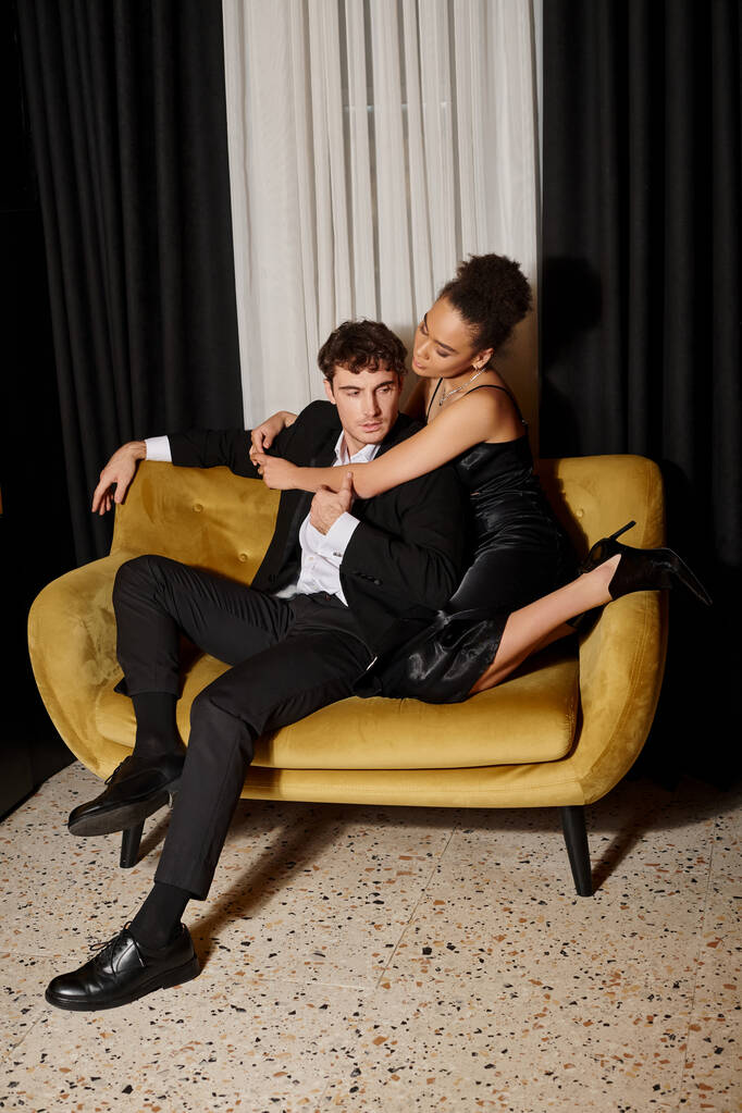 beautiful african american woman in dress embracing man in formal wear and posing on velvet sofa - Photo, Image
