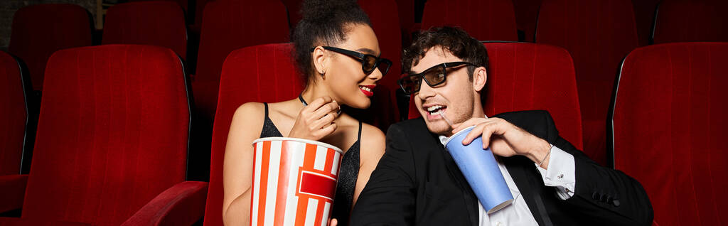 joyous diverse couple sitting on red cinema chairs and smiling at each other, Valentines day, banner - Photo, Image