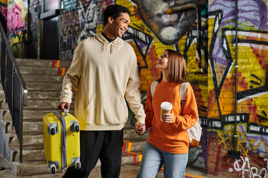 pareja diversa tomados de la mano y sonriendo con una maleta amarilla en una pared pintada de graffiti, albergue - Foto, Imagen