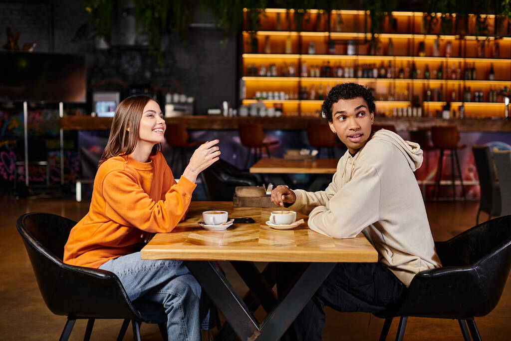 Χαρούμενη γυναίκα δείχνει κάτι σε μαύρο άνδρα, ενώ κάθονται μαζί σε ένα ζεστό καφέ, φλιτζάνια καφέ - Φωτογραφία, εικόνα