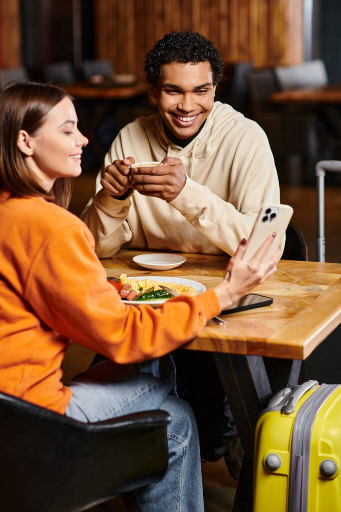 カラフルな服を着た笑顔の女性がスマートフォンを黒人のボーイフレンドにコーヒーカップで披露 - 写真・画像