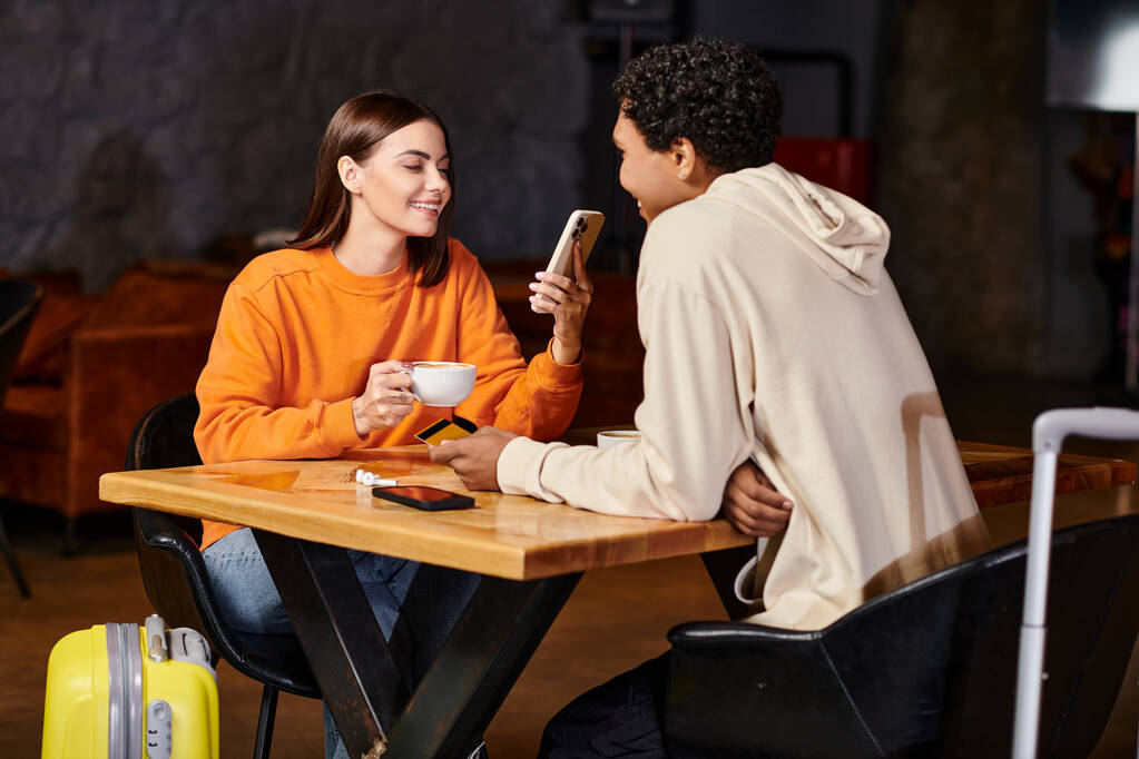 スマートフォンを使用して笑顔の若い女性とカフェで黒人のボーイフレンドの近くでコーヒーカップを保持 - 写真・画像