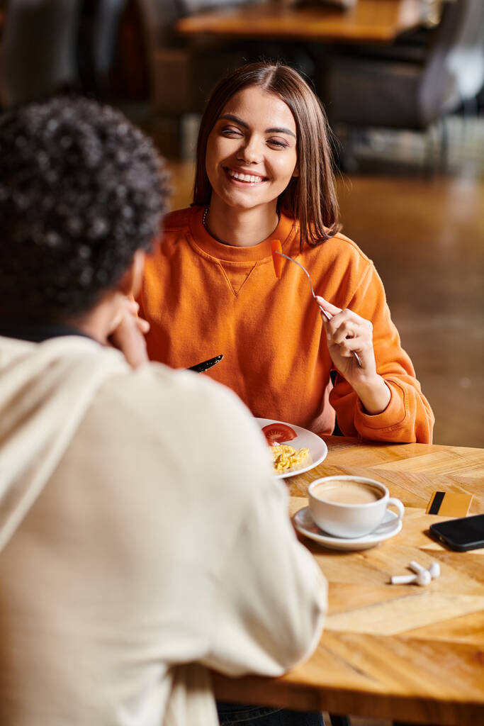 Glimlachende jonge vrouw heeft een heerlijk gesprek met vriendje tijdens een maaltijd in een gezellig café - Foto, afbeelding