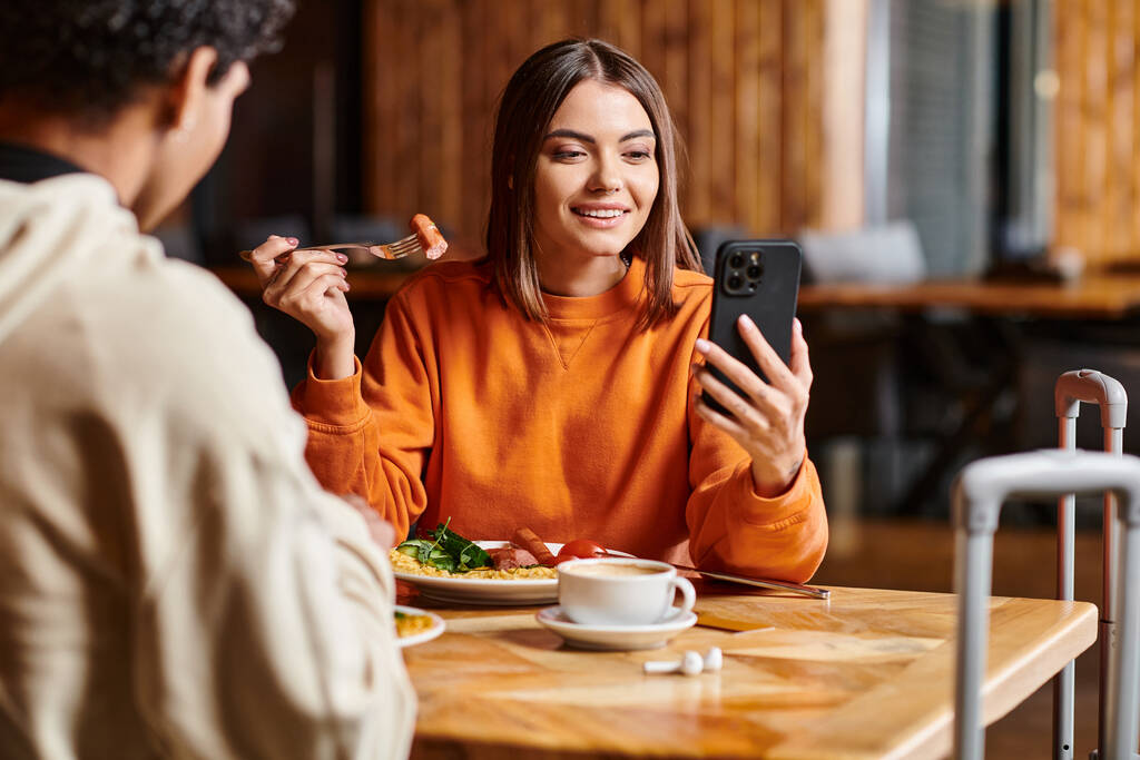 Nuori nainen elinvoimainen oranssi pusero iloisesti katsot hänen puhelimensa lähellä musta poikaystävä aterian aikana - Valokuva, kuva