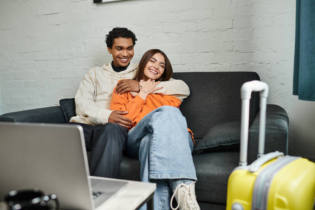 glückliches multiethnisches Paar kuschelt sich gemütlich auf einer Couch in ihrem stilvollen Wohnzimmer neben dem Laptop auf dem Tisch - Foto, Bild