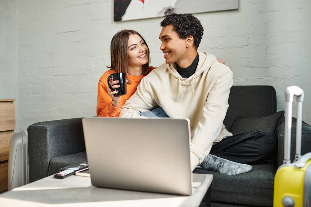 despreocupados pareja interracial comparte un momento acogedor mientras se sientan en el sofá y el uso de la computadora portátil, albergue - Foto, imagen