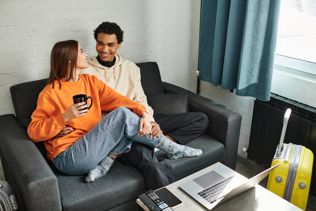 feliz casal diversificado em roupas aconchegantes sentado em um sofá no albergue, cercado por laptop e bagagem - Foto, Imagem