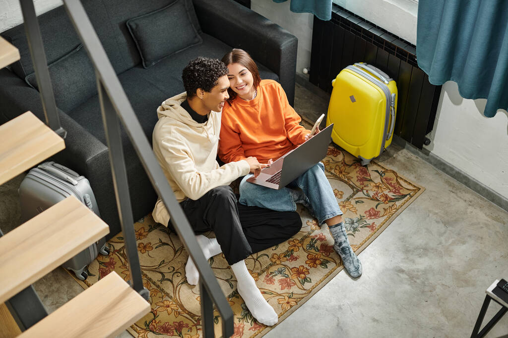 Smiling interracial couple sitting near sofa with laptop, planning a trip or discussing future plans - Photo, Image
