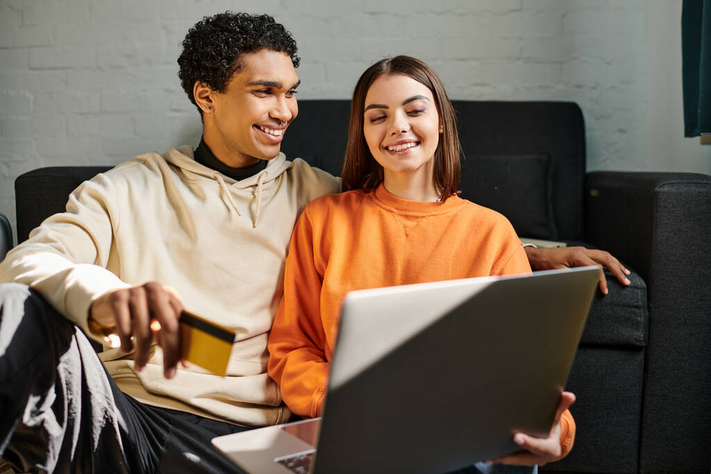 Diverso casal sorrindo compras on-line, segurando um cartão de crédito, com um laptop na sala de estar - Foto, Imagem