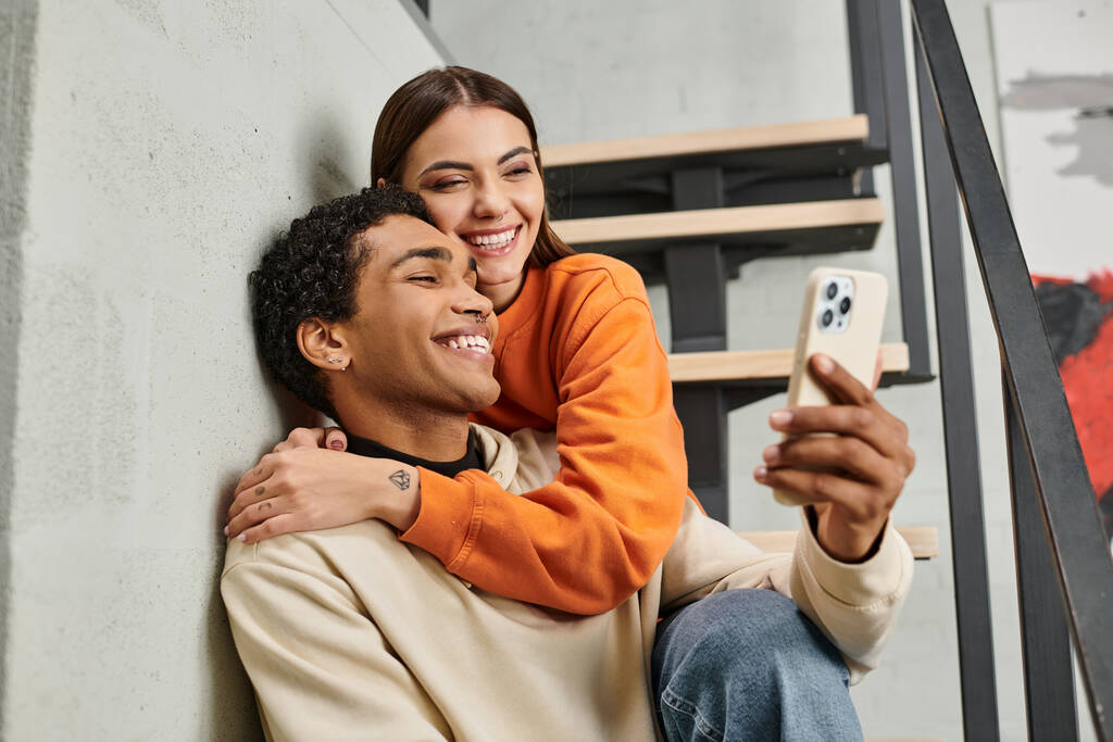 счастливая разнообразная пара сидит на лестнице и улыбается, делая селфи на смартфоне в общежитии - Фото, изображение