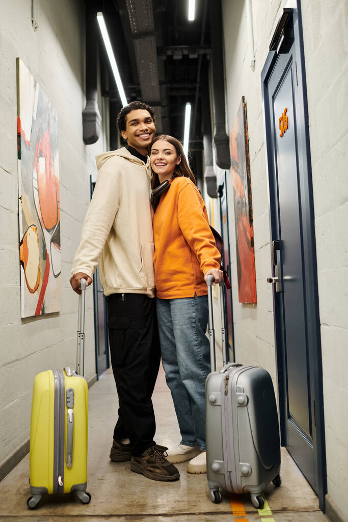 Feliz pareja diversa en un corredor de albergue con maletas, disfrutando de una experiencia de viaje alegre - Foto, Imagen
