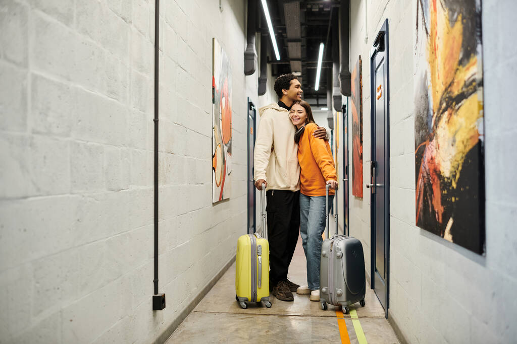 Coppia multiculturale rilassata con bagagli abbracciare e in piedi insieme in un corridoio ostello - Foto, immagini