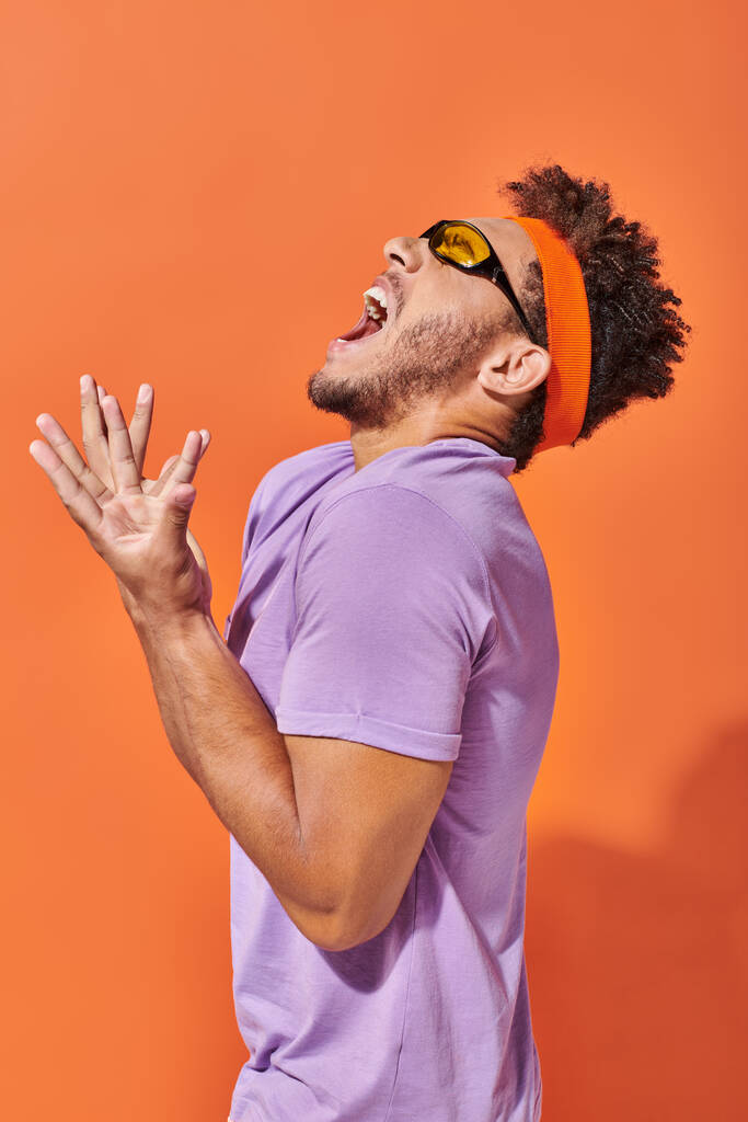 expressieve Afro-Amerikaanse man in bril en hoofdband schreeuwen op oranje achtergrond - Foto, afbeelding