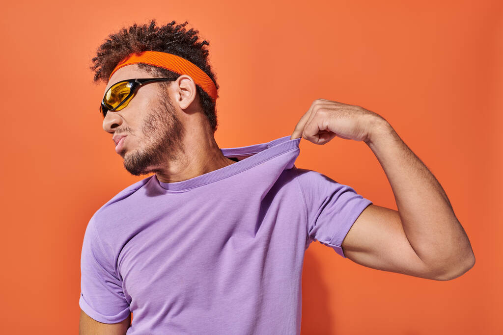 confiado hombre afroamericano en diadema ajustando camiseta púrpura sobre fondo naranja - Foto, Imagen