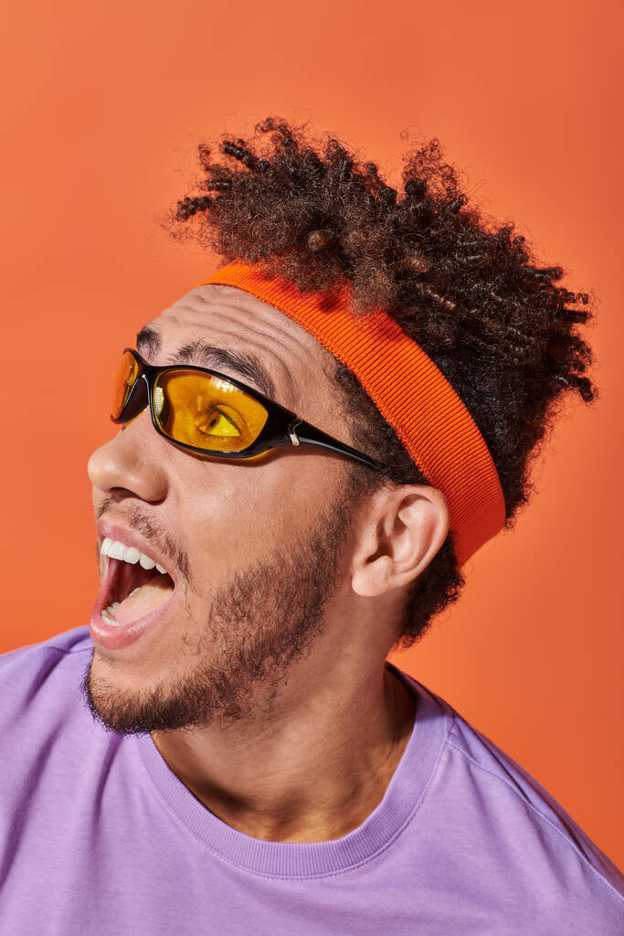 オレンジ色の背景に眼鏡とヘッドバンドで興奮するアフリカ系アメリカ人男性, 開いた口 - 写真・画像