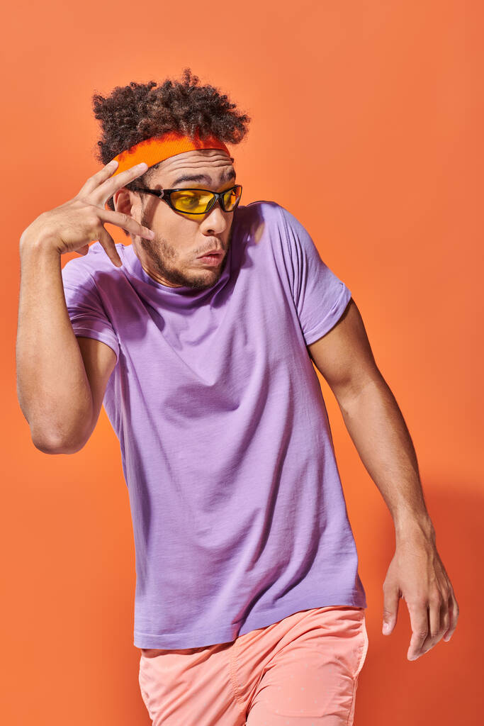 hombre afroamericano sorprendido en gafas y diadema mirando hacia otro lado sobre fondo naranja, gesto - Foto, Imagen
