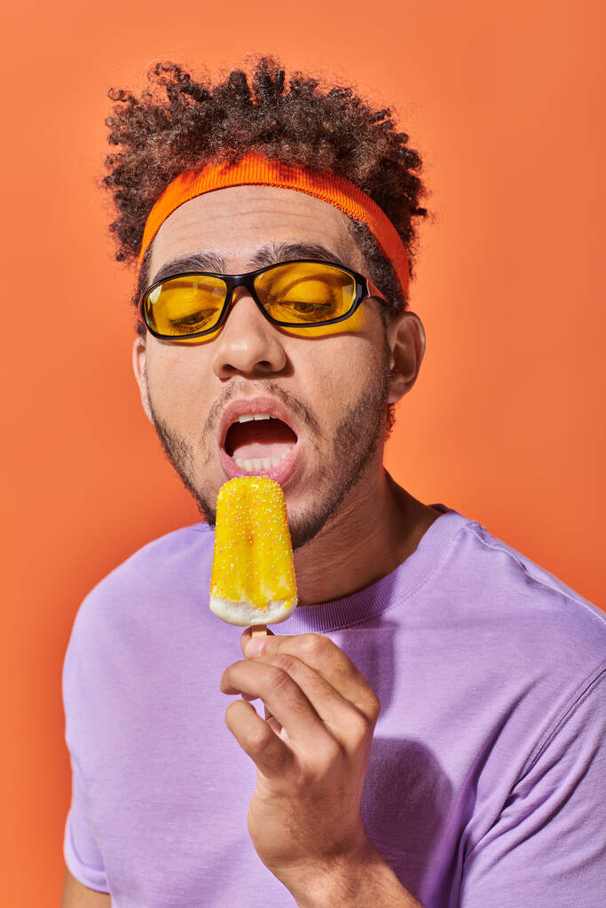 uomo afroamericano in occhiali da sole e cerchietto mangiare gelato fruttato in estate su sfondo arancione - Foto, immagini