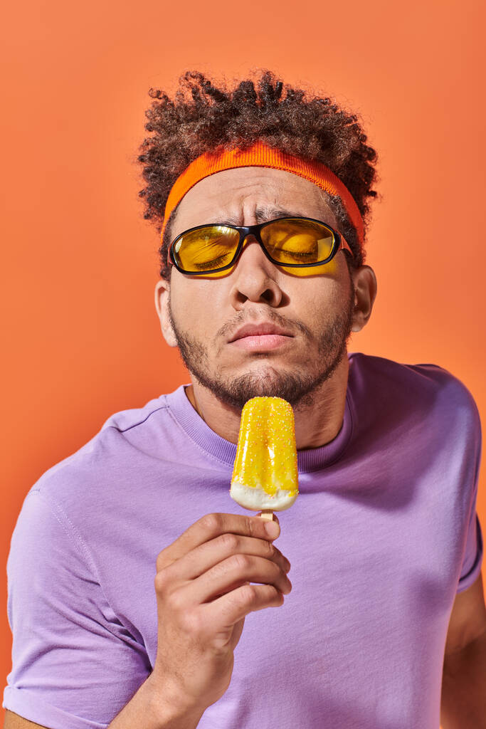 Африканский американец в солнцезащитных очках и ободок, нюхающий фруктовое мороженое на оранжевом фоне - Фото, изображение