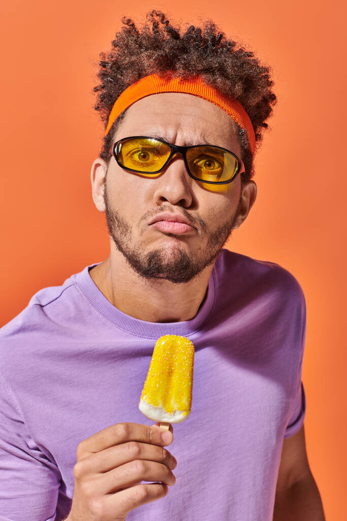 hombre afroamericano emocional en gafas de sol y diadema celebración de helado afrutado en naranja - Foto, imagen
