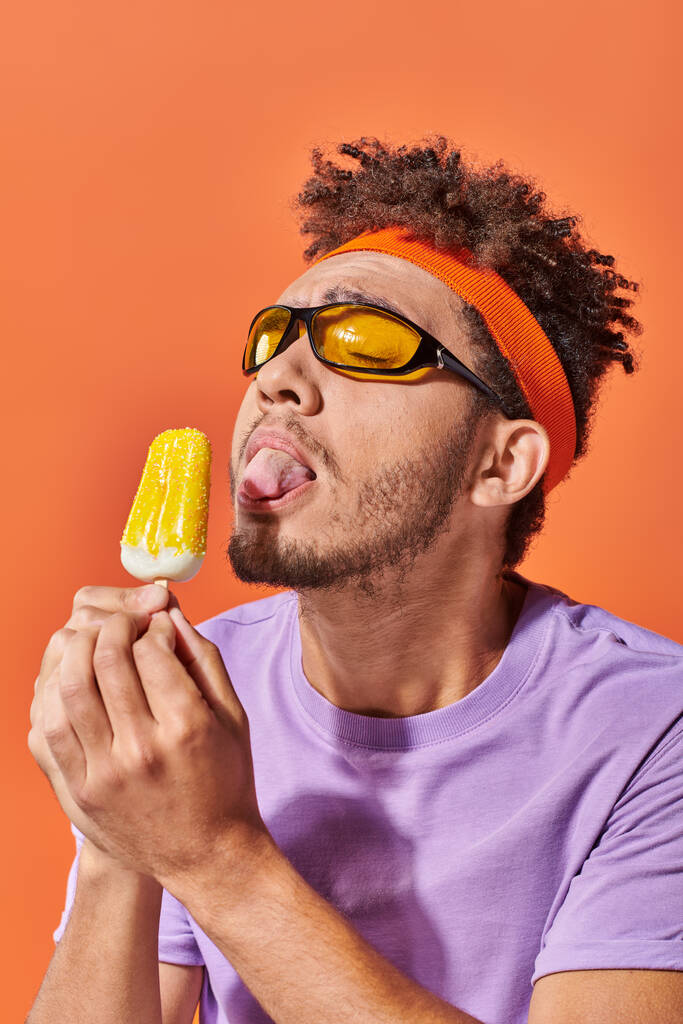 Afrikaan amerikaanse man in zonnebril en hoofdband likken fruitig ijs op oranje achtergrond, tong - Foto, afbeelding