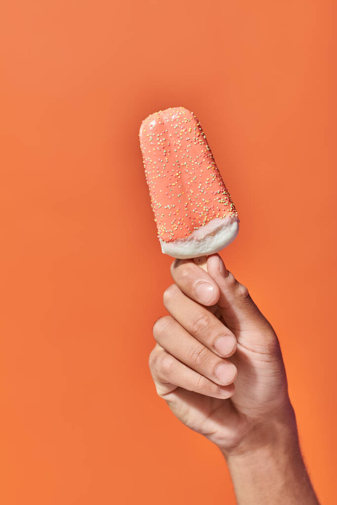 colpo ritagliato di persona che tiene congelato ghiacciolo gelato con spruzzi su sfondo arancione - Foto, immagini