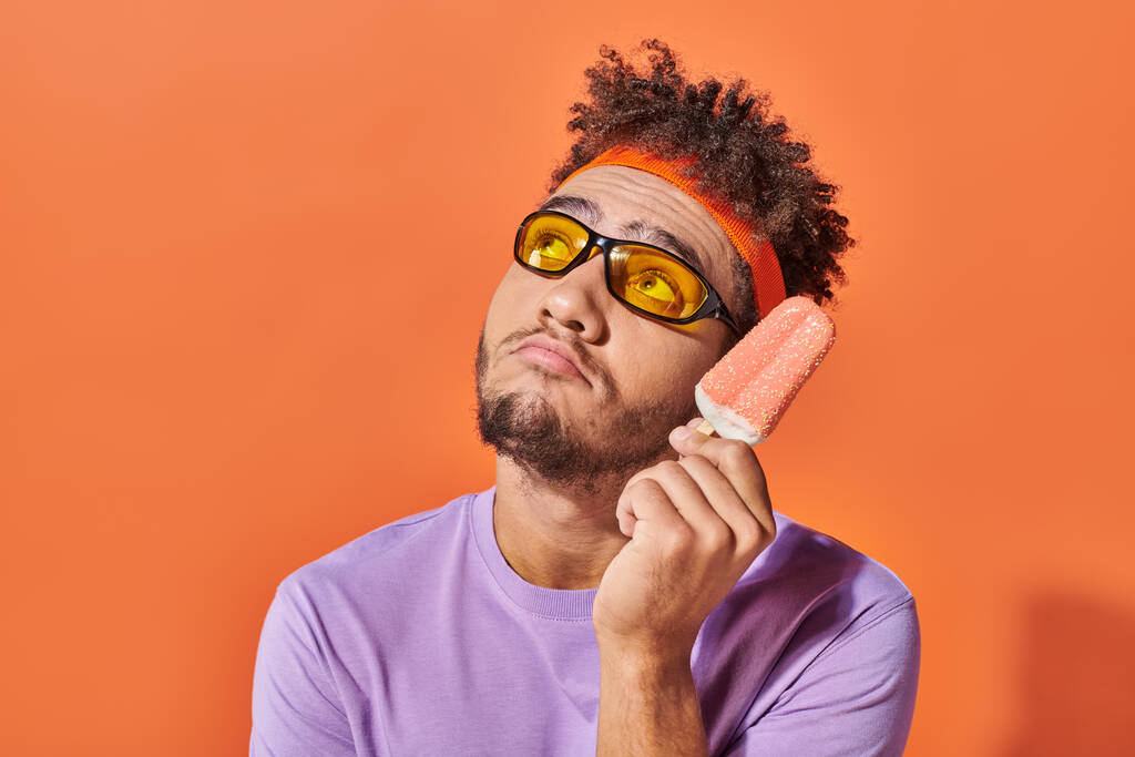 hombre afroamericano pensativo en gafas de sol celebración de helado con aspersiones sobre fondo naranja - Foto, imagen