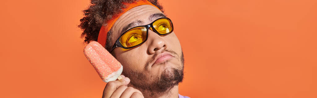 hombre afroamericano pensativo en gafas de sol celebración de helado con aspersiones en naranja, pancarta - Foto, Imagen