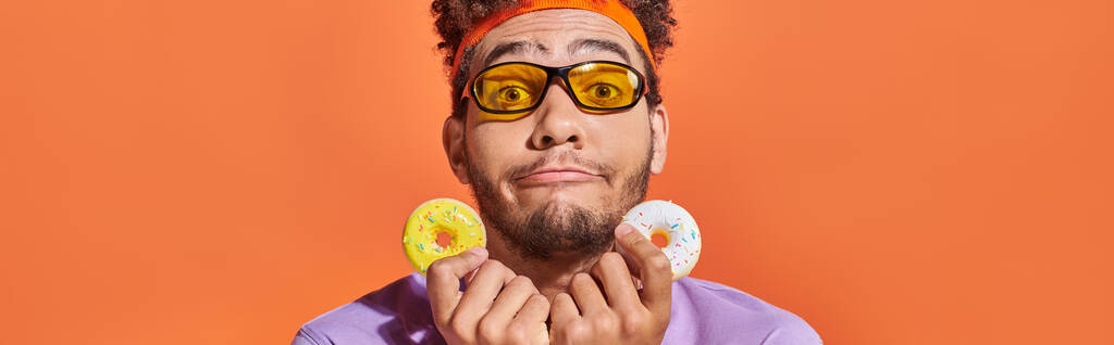 srandovní africký Američan v slunečních brýlích drží kousnutí velikosti koblihy na oranžovém pozadí, banner - Fotografie, Obrázek