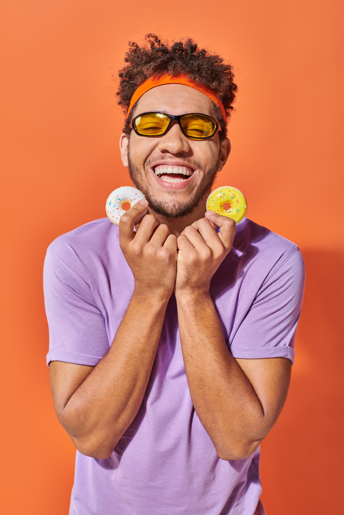 fröhlicher junger afrikanisch-amerikanischer Mann mit Sonnenbrille und mundgerechten Donuts auf orangefarbenem Hintergrund - Foto, Bild
