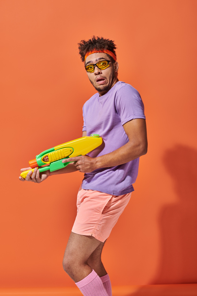 オレンジ色の背景にウォーターガンを保持するサングラスで混乱したアフリカ系アメリカ人男性, グリムス - 写真・画像
