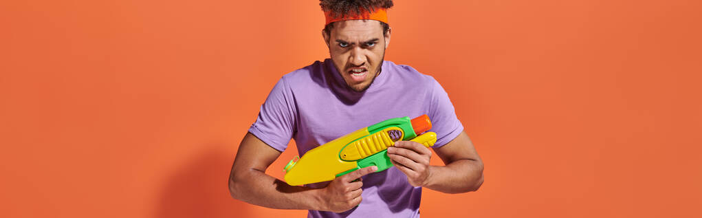 uomo afroamericano in fascia giocando lotta acqua con pistola giocattolo su sfondo arancione, banner - Foto, immagini