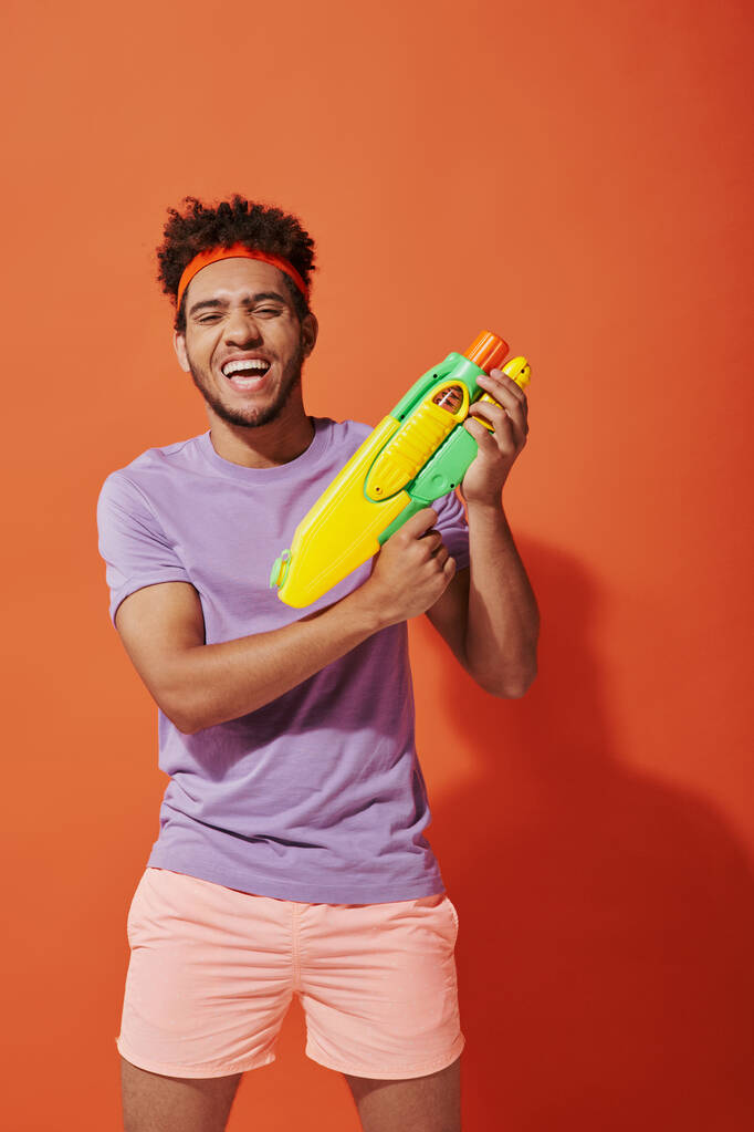 オレンジ色の背景におもちゃ銃で水を遊ぶヘッドバンドのポジティブなアフリカ系アメリカ人男性 - 写真・画像